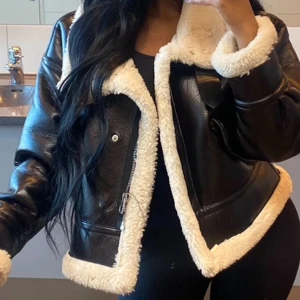 Svart jacka med fuskpäls från Zara - Säljer en svart skinnjacka från Zara med vit fuskpälsfoder och detaljer. Jackan har dragkedja, bälte i vid halsen och stora slag. Perfekt för kyliga dagar. Jag har storlek 36/S i överdel men för att få en liten oversized look så tog jag en storlek större. Se hur den sitter i första bilden. Använt ett par gånger, har en fläck vid krage går att tvätta bort, övrigt inga andra fel.