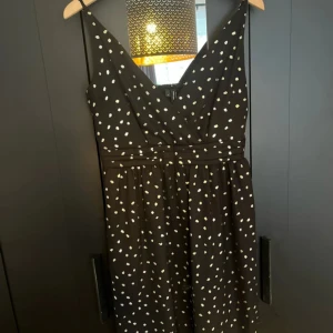 Svart prickig klänning med v-ringning - Svart klänning med vita prickar och v-ringad hals. Klänningen har breda axelband och markerad midja för en smickrande siluett. Perfekt för dig som gillar klassiska mönster med en modern twist.