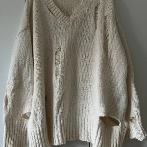 Beige stickad tröja med v-ringning - Säljer en oversized beige stickad tröja med v-ringning och slitna detaljer. Tröjan har långa ärmar och grovstickad struktur, perfekt för en avslappnad och trendig look.