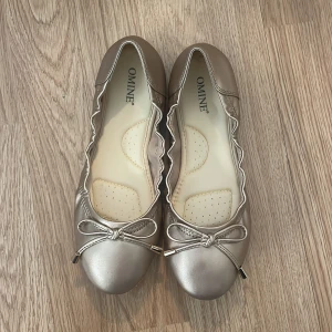 Silvriga ballerinaskor med rosett från Omine - Snygga populära silvriga ballerinaskor från Omine med rund tå och dekorativ rosett framtill. Skorna är helt oanvända.
