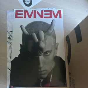 Eminem affisch - Säljer en stor affisch med Eminem. Affischen har en svartvit bild på Eminem som gör ett handtecken mot kameran och hans namn står i rött högst upp. Perfekt för dig som gillar hiphop och vill ha något snyggt på väggen.