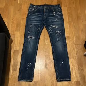 Dsquared2 - Mörkblå jeans från Dsquared2 med slitningar, färgstänk och en röd etikett vid fickan. Bakfickan har ett lönnlövsmärke och röda sömmar. Klassisk femficksmodell med knappgylf och skön passform.