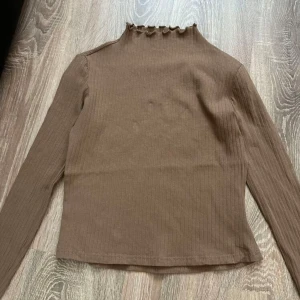 Beige ribbad långärmad topp med volangkant - Säljer en beige ribbad långärmad topp med volangkant vid halsringningen. Toppen har en smal passform och är tillverkad i ett mjukt, stretchigt material. Perfekt att styla med jeans eller kjol.