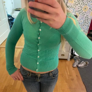 Grön kofta med knappar - Säljer en ljusgrön kofta med långa ärmar och vita knappar hela vägen framtill. Koftan är tight i modellen och har ribbade muddar vid ärmslut och nederkant. Perfekt att styla till jeans eller kjol.söljer då den inte passar längre