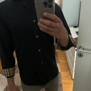 Svart skjorta från Burberry  - Säljer en svart skjorta från Burberry med klassiska beige, röda och svarta rutiga detaljer på insidan av kragen och ärmsluten. Skjortan är endast använd en gång på en skolavslutning där av skicket 10/10. Hör av er för eventulla frågor!!