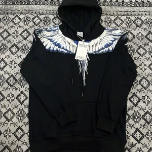 Svart hoodie med blå och vita vingar från Marcelo Burlon - Säljer en svart hoodie från Marcelo Burlon med ikoniskt vingtryck i blått och vitt över axlar och rygg. Tröjan har huva med dragsko och ribbade muddar. Perfekt för dig som vill sticka ut med en unik streetwear-look.
