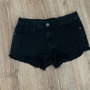 Svarta jeansshorts från UNE D - Svarta jeansshorts med fransad kant och klassisk femficksdesign. Shortsen har bälteshällor och knappstängning framtill. Perfekta för varma dagar och enkel att matcha med olika toppar.
