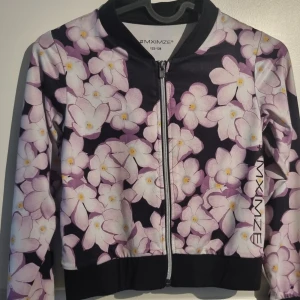 Blommig träningsjacka från MXIMZE - Snygg bomberjacka från MXIMZE med blommigt mönster i lila och vitt på mörk botten. Jackan har dragkedja framtill och svarta muddar vid ärmslut och midja. Perfekt för dig som vill sticka ut med färg och mönster!