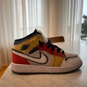 Nike Air Jordan 1 Mid multicolor - Säljer ett par Nike Air Jordan 1 Mid med färgblock i rött, gult, blått, svart och vitt. Skorna har svart snörning, vit sula och klassisk Jordan-logga på sidan. Perfekt för dig som vill sticka ut med dina sneakers.