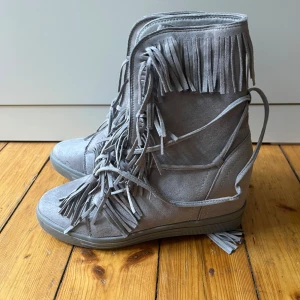 Grå mockaboots med fransar från Siader - Säljer ett par gråa fransskor. Skorna har en platt sula och rund tå med klack. Perfekta för sommaren! Storlek 38 men passar på 37 och 39.