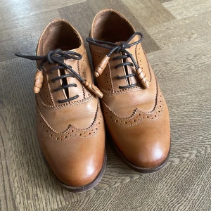 Bruna brogues från Massimo Dutti - Snygga bruna brogues från Massimo Dutti med klassiska perforerade detaljer och svarta snören med träpärlor. Skorna har låg klack och rund tå, perfekta för en stilren look. Tillverkade i skinn med fina sömmar och elegant design.