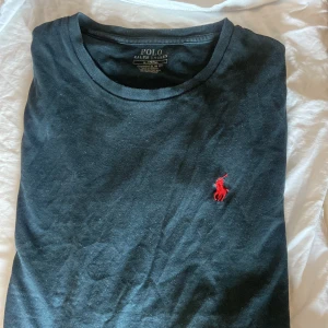 Svart t-shirt från Polo Ralph Lauren - Svart t-shirt från Polo Ralph Lauren med klassisk rund hals och röd broderad logga på bröstet. Modellen är custom slim fit och tillverkad i mjuk bomull. Perfekt för en stilren och enkel look.