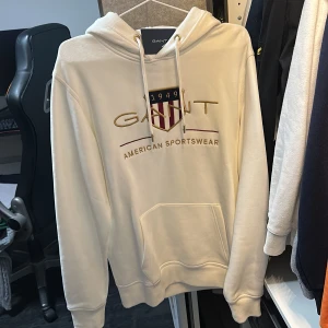 Vit hoodie från GANT med broderad logga - Säljer en vit hoodie från GANT med broderad logga och texten 'American Sportswear' framtill. Tröjan har huva med dragsko och en stor magficka. Perfekt för en avslappnad och sportig stil.