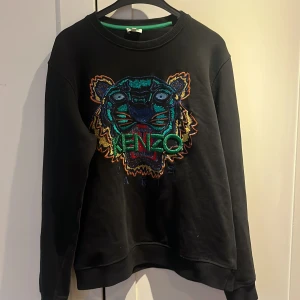 Svart sweatshirt med tigerbrodyr från Kenzo - Cool svart sweatshirt från Kenzo med ikonisk tigerbrodyr i färgglada nyanser och Kenzo-logga framtill. Tröjan har rund halsringning och långa ärmar. Perfekt statement-plagg för dig som gillar streetwear och vill sticka ut.