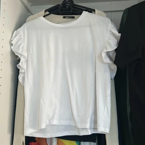 Vit topp med volangärmar från Zara - Säljer en vit topp från zara med rund halsringning och volangdetaljer på ärmarna. Perfekt för en enkel men söt look. Tunn och skön bomullskvalitet. Har använt 1-2 gånger.  Pris kan diskuteras 
