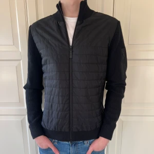 Svart hybridjacka från J.Lindeberg - Snygg svart hybridjacka från J.Lindeberg med quiltad front och stickade ärmar och rygg. Jackan har dragkedja framtill, ståkrage och en praktisk ficka med dragkedja på ärmen. I princip oanvänd. Nypris 3200kr. 