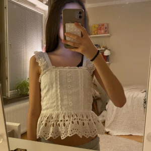 Vit broderad linnetopp med volang - Söt vit linnetopp med breda axelband och volangkant nertill. Toppen har broderade detaljer och hålmönster, samt volang på axelbanden. 