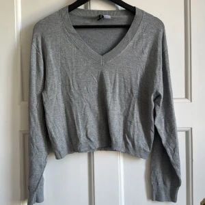 Grå croppad v-ringad tröja från Divided - Säljer en grå croppad tröja från Divided med v-ringning och långa ärmar. Perfekt för lager-på-lager och enkel att matcha med olika outfits. Mjuk och bekväm med ribbade muddar vid ärmslut och hals.