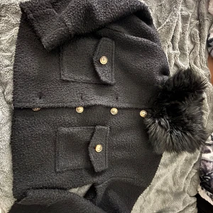 Svart kappa med fuskpäls och guldknappar - Säljer en svart kappa med stora guldknappar och två fickor framtill. Jackan har en krage och dekorativ fuskpäls vid ärmsluten. Materialet är grovt och ger en lyxig känsla. Perfekt för kyligare dagar. Köpt fårn Chelsea och kappan går för 800-1000kr, passar både S och XS