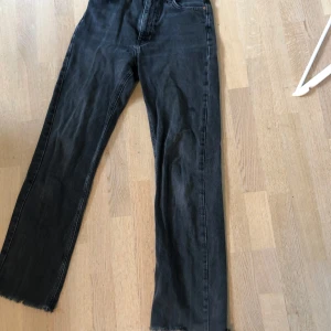 Mörkgrå jeans från NA-KD - Säljer ett par mörkgrå jeans från NA-KD med raka ben och råa benslut. Klassisk femficksmodell med knapp och dragkedja framtill. Ser mörkare ut på långdistansbild, är en mer realistisk färg på närbilden.