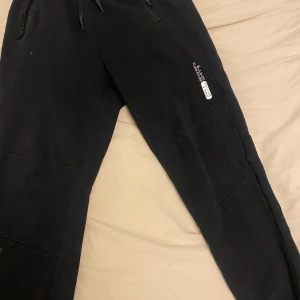 Svarta mjukisbyxor med dragkedjor - Svarta mjukisbyxor med dragsko i midjan och dragkedjor vid fickorna. Byxorna har en liten vit logga på ena benet och är perfekta för en avslappnad stil.