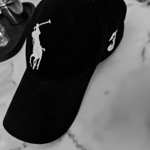 Svart keps från Polo Ralph Lauren - Svart keps med stor vit broderad polospelare framtill och siffran 3 på sidan. Baktill står det 'POLO' broderat i vitt. Klassisk böjd skärm och justerbar passform. Perfekt accessoar för en sportig och stilren look.