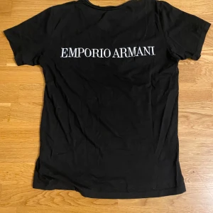Svart t-shirt från Emporio Armani - Säljer en svart t-shirt från Emporio Armani med vit logga på bröstet och stor vit text på ryggen. Klassisk passform och rund hals. Perfekt för dig som gillar stilrena märkesplagg.