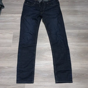 Mörkblå jeansbyxor från Dressmann - Säljer ett par klassiska mörkblå jeansbyxor från Dressmann med raka ben och kontrastsömmar. Byxorna har fem fickor och stängs med dragkedja och knapp. Perfekta för dig som gillar enkel och stilren look.