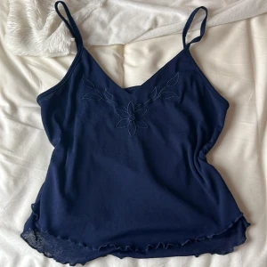 Mörkblå linnetopp med broderad blomma - Säljer en mörkblå linnetopp med smala axelband och broderad blomma framtill. Toppen har volangkant nertill och är perfekt för varma dagar.