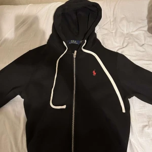  Ralph Lauren hoodie  - Svart hoodie från Polo Ralph Lauren med dragkedja framtill och vit dragsko i huvan. Klassisk röd logga broderad på bröstet. Perfekt för en avslappnad och stilren look