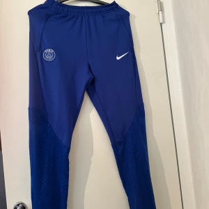 Blå Nike PSG träningsbyxor - Säljer ett par blå träningsbyxor från Nike med PSG-logga på ena benet och Nike-logga på andra. Byxorna har slim fit-passform, resår i midjan och är tillverkade i ett syntetmaterial som andas. Perfekta för fotbollsträning.