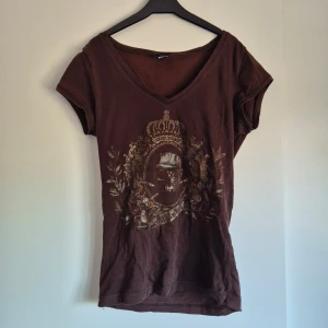 Brun t-shirt med guldtryck och v-ringning - Säljer en brun t-shirt med v-ringad hals och korta ärmar. Framsidan har ett detaljerat guldmönster med en dödskalle och dekoration. Perfekt för dig som gillar unika tryck. Y2K