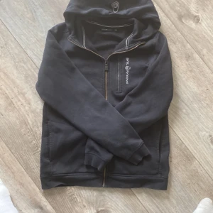 Svart hoodiejacka från Sail Racing - Svart hoodiejacka från Sail Racing med dragkedja framtill och en extra bröstficka med dragkedja. Jackan har broderad logga på bröstet och mjukt fleecefoder på insidan. Perfekt för dig som gillar sportig stil.
