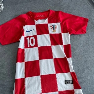 Kroatien fotbollströja Nike Modric 10 - Säljer en Kroatien landslagströja från Nike med Modric och nummer 10. Tröjan har klassiskt rödvitt rutmönster, blå Nike-logga och landslagsemblem på bröstet. Korta ärmar och tillverkad i lätt, ventilerande material.