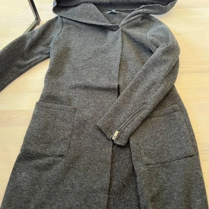 Grå lång hoodie-kappa från Amisu - Mysig grå kappa med huva från Amisu. Kappan har stora fickor framtill och långa ärmar. Perfekt för lager-på-lager och enkel att matcha med olika outfits.