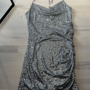 Silver paljettklänning från BikBok - Säljer en glittrig silverfärgad paljettklänning från BikBok med halterneck och draperad detalj på sidan. Klänningen är kort och har en snygg omlottkänsla framtill. Perfekt för dig som vill sticka ut! Använd en gång. 