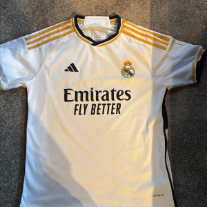 Real Madrid fotbollströja Bellingham 5 - Säljer en vit Real Madrid matchtröja från Adidas med Bellingham 5 på ryggen. Tröjan har korta ärmar, svarta och gula detaljer samt klubbmärke och sponsortryck på bröstet. Slim fit-modell, perfekt för fans! Använd max 10 gånger. 10/10 skick