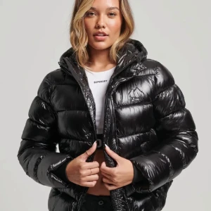 Svart glansig pufferjacka från Superdry - Säljer en svart, glansig pufferjacka från Superdry med huva och dragkedja. Superfint skick utöver ett litet hål på ena armen som är väldigt lättfixat (kan laga innan jag skickar om så önskas)❤️Hör av er vid frågor❤️