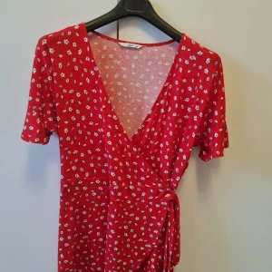 Omlottstopp från Ellos strl 46/48 - Röd omlottstopp från Ellos med små vita blommor och knyt i midjan. Klänningen har v-ringning och korta ärmar. Tillverkad i mjuk viskosblandning med lite stretch för extra komfort. Storlek 46/48. Kan hämtas i Vallentuna.