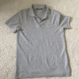 Grå pikétröja från J.Lindeberg - Säljer en stilren grå pikétröja från J.Lindeberg. Tröjan har klassisk krage, korta ärmar och två knappar framtill. Diskret logga på bröstet. Sitter tight vid armarna. 