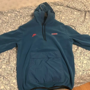 Blå fleece hoodie från Nike Air Max - Säljer en blå hoodie från Nike Air Max med broderad logga och Air Max-märke på bröstet. Tröjan har huva med dragsko och stor magficka framtill. Tillverkad i mjuk fleece, perfekt för kyliga dagar.