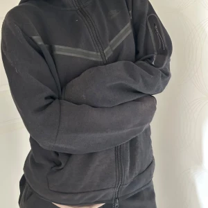 Svart zip-hoodie och sweatpants från Nike - Svart hoodie och swetpants från Nike med dragkedja framtill och diskreta svarta detaljer över bröstet och benen. Jackan har lång ärm och huva, samt två fickor på sidorna. Två djupa fickor på sidan av benen Perfekt för en sportig och avslappnad stil.
