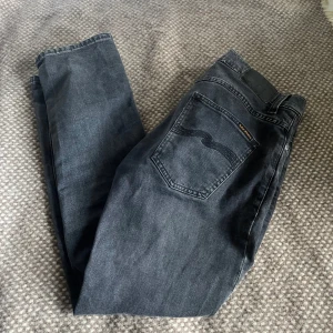 Nudie Grim Tim Mörkgråa Jeans - Mörkgrå Nudie Jeans i modellen Grim Tim, storlek W32 L30. Jeansen är i mycket fint skick, med snygg slim passform och bekväm stretch. Ett tidlöst basplagg i hög kvalitet – perfekta för både vardag och mer uppklädda tillfällen. Grim Tim-modellen är rakare i passformen och funkar lika bra till sneakers som boots. 