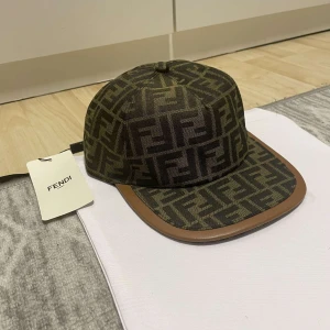 Fendi keps med monogram - Snygg keps från Fendi med klassiskt FF-monogram i brunt och svart. Skärmen har en brun kant i läderimitation och kepsen har justerbar rem baktill. Perfekt accessoar för att lyfta din outfit.