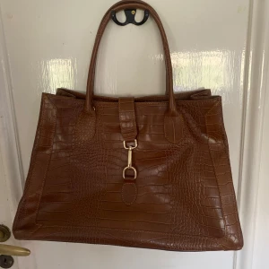 Brun krokopräglad handväska - Säljer en snygg brun handväska med krokodilpräglat mönster!  Väskan är 42 cm bred och 28 cm lång. Den kommer även med ett längre band man kan sätta på. Skriv om ni vill ha mer bilder eller frågor!❤️❤️