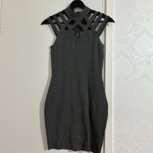 Grå tight klänning med unikt axelparti -  grå tight klänning med höghalsad krage och coola banddetaljer över axlarna. Klänningen är ärmlös och har dragkedja med pärldetalj i nacken. Perfekt för dig som gillar en stilren men edgy look.