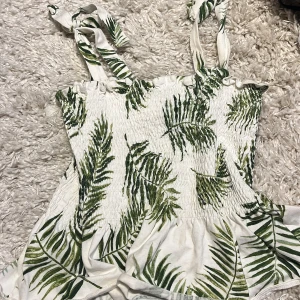 Vit sommarblus med grönt blad-mönster från H&M - Supersöt vit blus från H&M med gröna blad och smock upptill. Axelbanden är knytbara och blusen har en luftig passform, perfekt för varma dagar. Säljer då den inte kommer till användning längre!