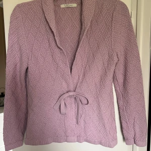 Ljuslila stickad kofta från First Class - Aldrig använd! Inköpt på gekås 129:-. Mysig ljuslila stickad kofta från First Class med knytband i midjan och lång ärm. Koftan har en mjuk struktur och en öppen design framtill. Perfekt för lager-på-lager och enkel att styla till olika outfits.