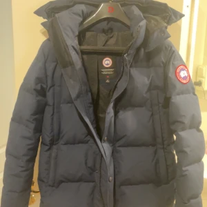 Mörkblå dunjacka från Canada Goose - Säljer en mörkblå Wyndham Core Reset dunjacka från Canada Goose med klassisk patch på ärmen. Jackan har hög krage, huva och dragkedja med knappar framtill. Perfekt för kalla vinterdagar och riktigt snygg streetstil. Fylld med dun och har flera praktiska fickor. Köpt från Johnells, kvitto kan visas upp. Nypris 15,999kr