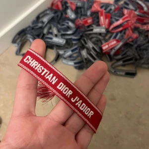 Rött Christian Dior J'Adior armband - Snyggt rött armband från Christian Dior med texten 'J'Adior' broderad i vitt. Armbandet har justerbar knytning med röda och vita tofsar i ändarna. Perfekt accessoar för att lyfta din outfit med en lyxig touch.
